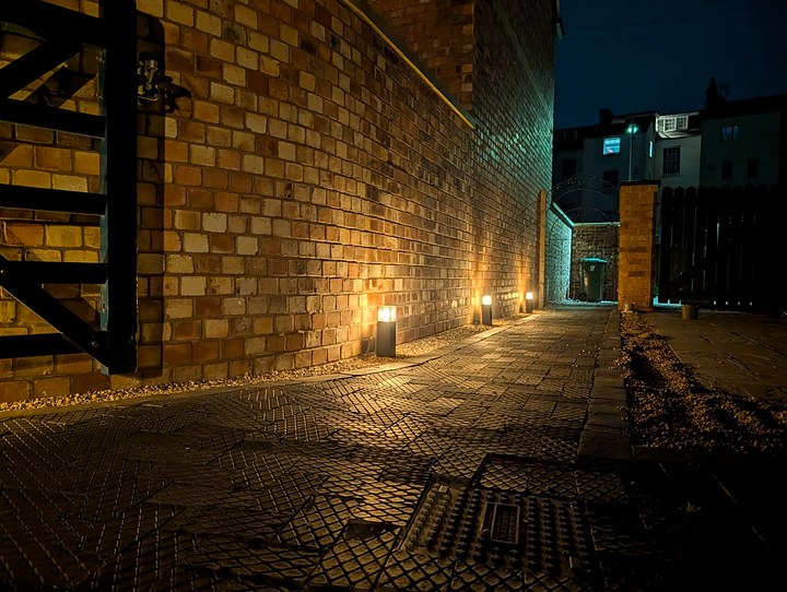 Courtyard path at night with low bollard lights glowing along a tall brick wall