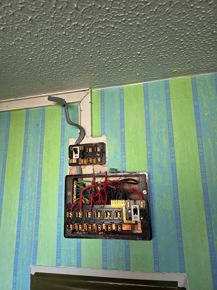 Old fuse box with exposed wiring on a striped wallpapered wall before a rewire